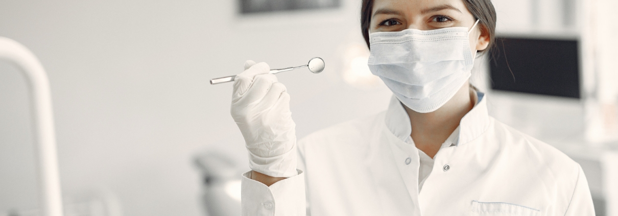 A female dentist wearing a white coat, gloves, and a face mask holds up a dental mirror in a dental office.