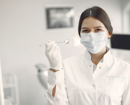 A female dentist wearing a white coat, gloves, and a face mask holds up a dental mirror in a dental office.