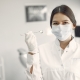 A female dentist wearing a white coat, gloves, and a face mask holds up a dental mirror in a dental office.