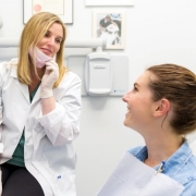 A dentist wearing a white coat and gloves talks to a smiling patient sitting in a dental chair, with certificates on the wall in the background.