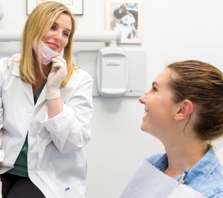 A dentist wearing a white coat and gloves talks to a smiling patient sitting in a dental chair, with certificates on the wall in the background.