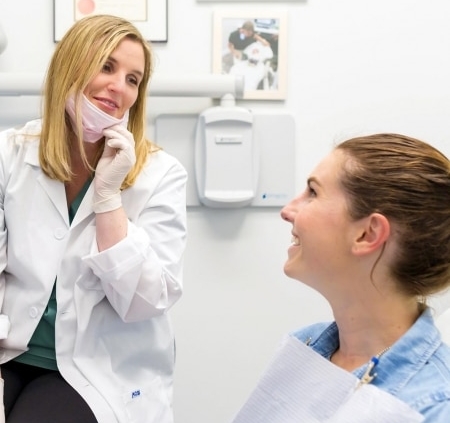 A dentist wearing a white coat and gloves talks to a smiling patient sitting in a dental chair, with certificates on the wall in the background.