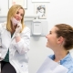 A dentist wearing a white coat and gloves talks to a smiling patient sitting in a dental chair, with certificates on the wall in the background.