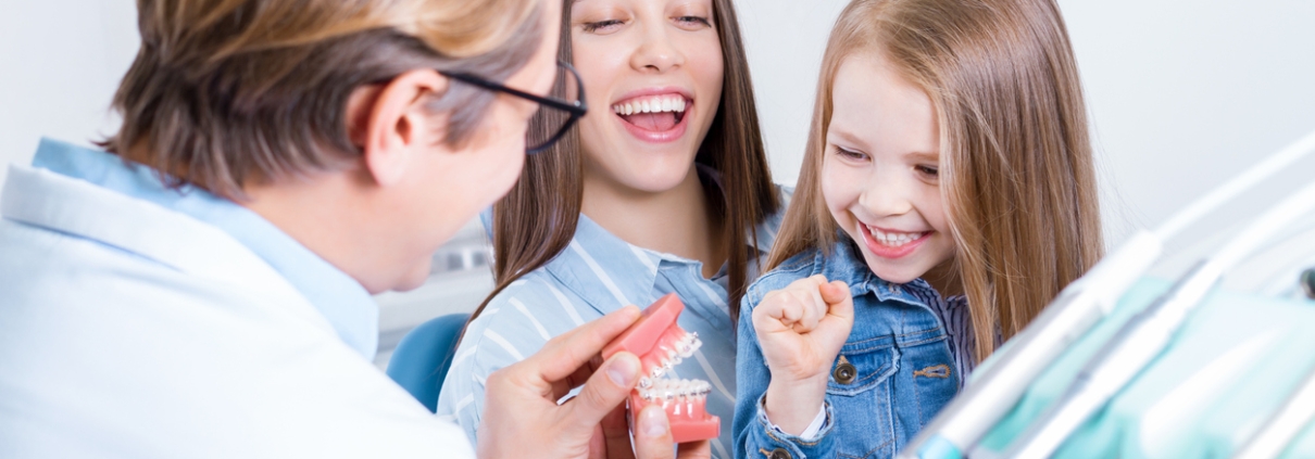 A dentist shows a dental model to a smiling young girl and a woman, likely her mother, in a dental office.