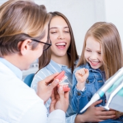 A dentist shows a dental model to a smiling young girl and a woman, likely her mother, in a dental office.