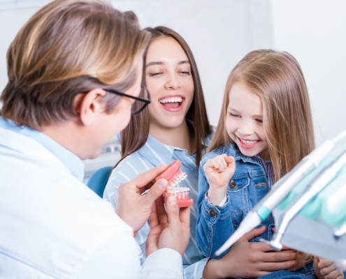 A dentist shows a dental model to a smiling young girl and a woman, likely her mother, in a dental office.