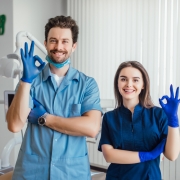 Two dental professionals wearing blue scrubs and gloves stand in a dental clinic, smiling and making an OK gesture with their hands.