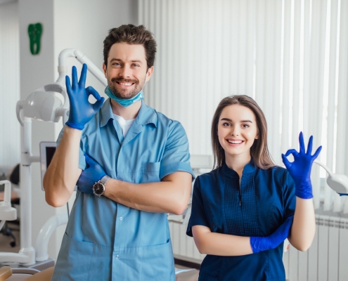 Two dental professionals wearing blue scrubs and gloves stand in a dental clinic, smiling and making an OK gesture with their hands.