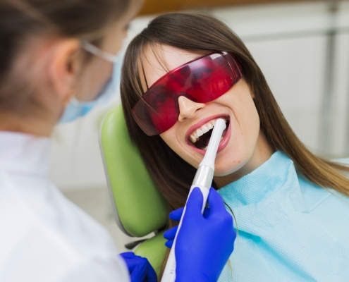 A dentist in gloves uses a dental tool to clean a womans teeth; the woman wears protective eyewear and a dental bib.