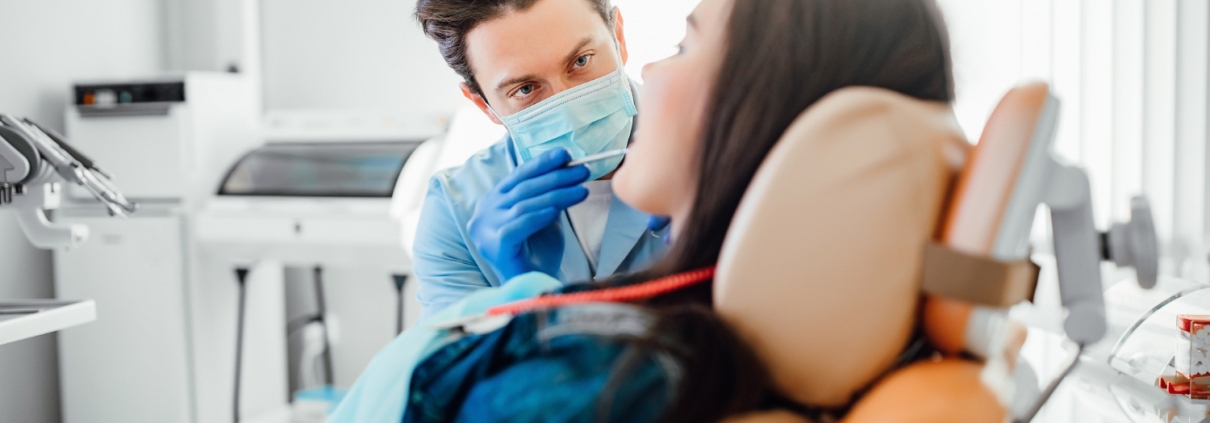A dentist wearing gloves and a mask examines a patient who is reclining in a dental chair in a brightly lit clinic.