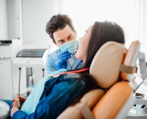 A dentist wearing gloves and a mask examines a patient who is reclining in a dental chair in a brightly lit clinic.